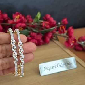 Puede incluir: Dos brazaletes dorados y plateados con un diseño de infinito repetido, incrustados con pequeñas piedras preciosas transparentes. Los brazaletes se sostienen en una mano, con un fondo borroso de flores rojas y una superficie de madera. También es visible una etiqueta blanca con el texto "Nsquare Collections".