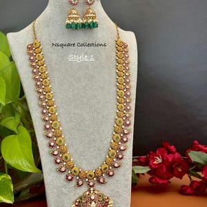 May include: A gold-toned necklace and matching jhumka earring set featuring red and green stones with white accents. The necklace has a long chain with circular motifs and a large pendant. The text Nsquare Collections Style-1 is visible.