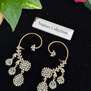 Op de afbeelding: Een paar goudkleurige oorbellen met een bloemmotief. Elke oorbel heeft een gebogen bovenkant, een centrale rechthoekige tealkleurige steen en hangende druppelvormige tealstenen, allemaal omringd door kleine, heldere kristallen. De oorbellen staan tegen een zwarte achtergrond.