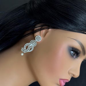 Pu&ograve; includere: Eleganti orecchini pendenti color argento con un design floreale e a spirale. Gli orecchini presentano piccole pietre scintillanti e una perla pendente. Gli orecchini sono esposti su un manichino.