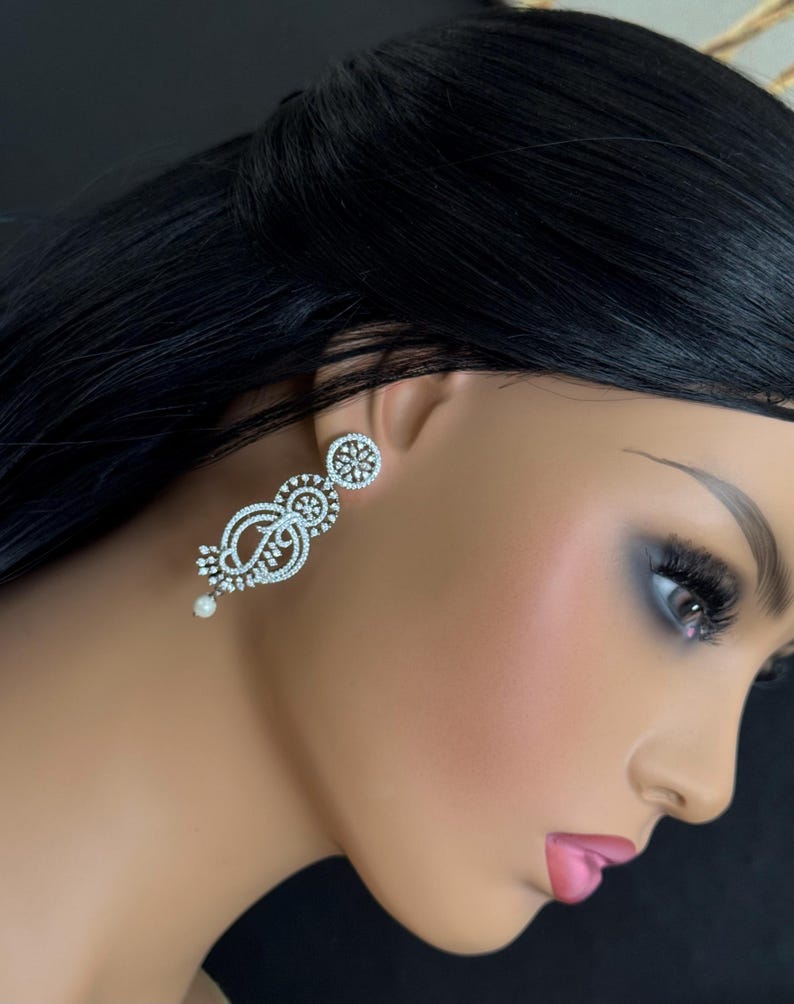 Pu&ograve; includere: Eleganti orecchini pendenti color argento con un design floreale e a spirale. Gli orecchini presentano piccole pietre scintillanti e una perla pendente. Gli orecchini sono esposti su un manichino.