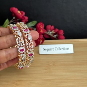 Può includere: Due braccialetti dorati con intricati disegni a filigrana, adornati con pietre preziose chiare e color rubino. I braccialetti sono esposti su uno sfondo di fiori rossi e una superficie di legno. Il testo "Nsquare Collections" è visibile.