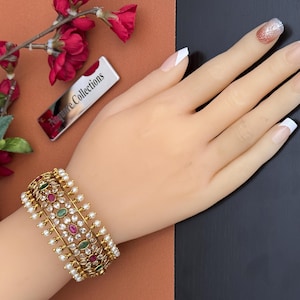 May include: A gold-toned bracelet with pearls, red and green gemstones. The bracelet is on a mannequin hand with French-tip nails. A small silver tag with the text "Aspire Collections" is visible.