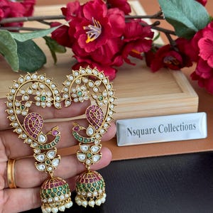 May include: Gold-toned earrings with a heart-shaped design, featuring white, green, and red gemstones. The earrings have a peacock motif and a bell-shaped bottom with small pearls. The text "Nsquare Collections" is visible.