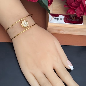 May include: A gold-toned double-band cuff bracelet featuring a central starburst charm encrusted with small clear stones. The bracelet has a smooth, polished finish and a minimalist open design that rests on the wrist.
