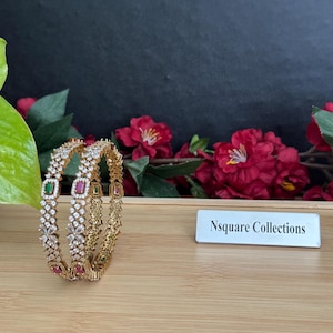 May include: Two gold-colored bangles with clear and colored gemstones. The bangles have a floral and geometric design, with green and red square-cut stones. The background includes red flowers and a wooden surface.