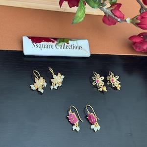 May include: Four pairs of gold-toned earrings with various designs. Two pairs feature floral and leaf motifs with clear and red gemstones. The earrings are displayed on a dark surface, with the text "Nsquare Collections" visible.