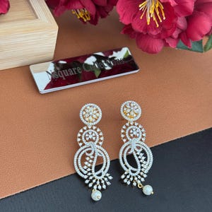 Pu&ograve; includere: Un paio di orecchini pendenti color oro con dettagli intricati di pietre bianche scintillanti. Ogni orecchino presenta una parte superiore floreale, tre disegni circolari e una base a forma di goccia con una piccola perla.