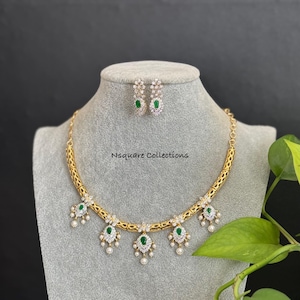 May include: A gold-coloured necklace and matching earrings set. The necklace features emerald-green gemstones and small white pearls. The earrings have a similar design with emerald-green gemstones. The text "Nsquare Collections" is visible.