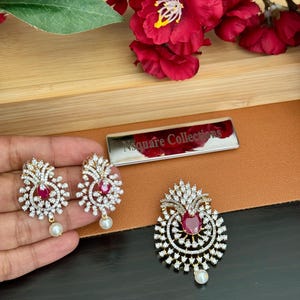 May include: A gold-coloured jewellery set comprising a pendant and matching earrings. Each piece features a central red gemstone surrounded by clear crystals. The earrings and pendant have a dangling white pearl. The brand name "Nsquare Collections" is visible.