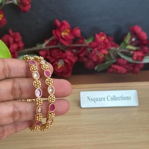 May include: Gold-toned bangles with oval-shaped pink and clear gemstones. The bangles are adorned with small, round gold accents. The background features red flowers and the text "Nsquare Collections".