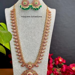 May include: A gold-toned necklace and earring set displayed on a mannequin. The necklace features a series of red and white gemstone accents. The earrings and necklace have green bead accents. The text "Nsquare Collections" is visible.
