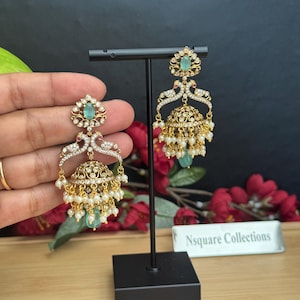 May include: Gold chandelier earrings with a floral design and dangling pearls. The earrings feature a central emerald-green stone and are displayed on a black stand. The earrings have a detailed, ornate design.