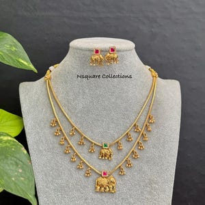 Può includere: Un set composto da una collana a due fili e orecchini a lobo abbinati color oro. La collana presenta piccoli ciondoli a forma di campanella e due pendenti a forma di elefante, uno con una pietra quadrata verde e l'altro con una pietra quadrata rossa. Il testo Nsquare Collections è visibile.