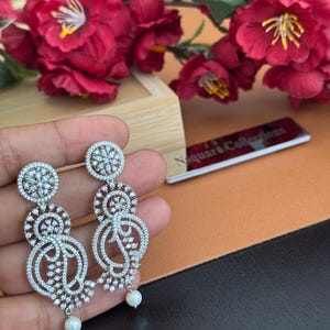 Pu&ograve; includere: Un paio di orecchini pendenti color argento con intricati disegni traforati e piccole perle bianche rotonde sul fondo. Gli orecchini sono adornati da numerosi piccoli cristalli trasparenti. Lo sfondo include fiori rossi e un biglietto da visita.