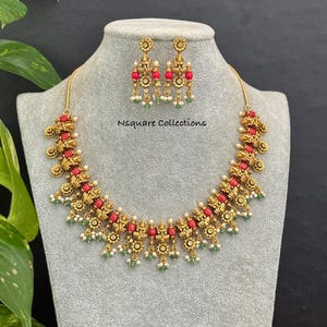 May include: A gold-toned necklace and matching earring set featuring intricate metalwork, red beads, and small green and white accents. The necklace has a central pendant design with repeating floral motifs and the text Nsquare Collections.