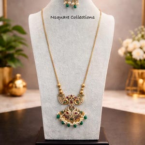 May include: Gold-toned necklace and earring set displayed on a mannequin. The necklace features a pendant with red, green, and white accents, and the earrings have a similar design. The set is adorned with small pearls and green gemstones. The text "Nsquare Collections" is visible.