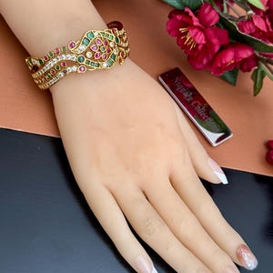 May include: A gold-toned cuff bracelet featuring an intricate openwork design adorned with embedded red, green, and clear stones. The bracelet displays a symmetrical pattern of floral and leaf-like motifs, showcasing a polished metallic finish.