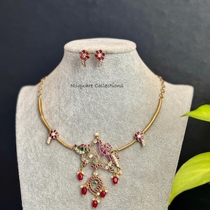 May include: Gold-toned necklace and matching earrings set. The necklace features a pendant with a bird design, red and green gemstones, and dangling red beads. The earrings are small, flower-shaped, and red. The text "Nsquare Collections" is visible.