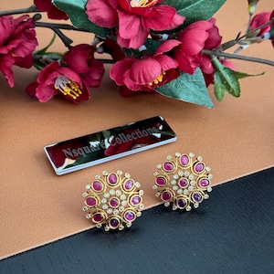 May include: A pair of gold-toned stud earrings with a circular design. Each earring features a central cluster of red gemstones surrounded by smaller clear stones. The earrings are set against a brown and black background, with a floral arrangement in the background.