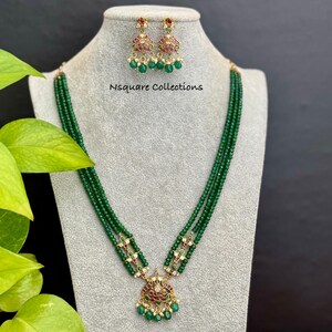 May include: A green beaded necklace and matching earrings set displayed on a mannequin. The necklace has a pendant with red and white accents. The earrings and pendant have a gold-colored setting. The text "Nsquare Collections" is visible.