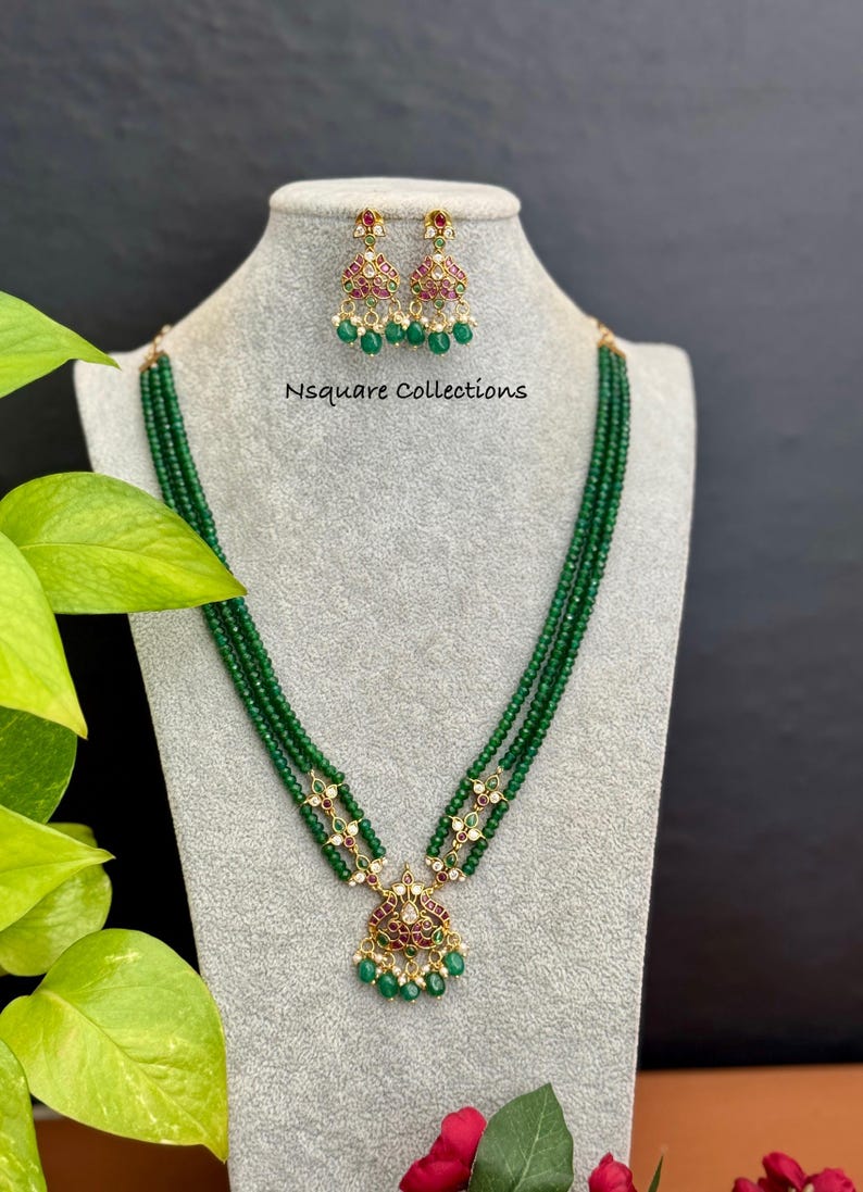 May include: A green beaded necklace and matching earrings set displayed on a mannequin. The necklace has a pendant with red and white accents. The earrings and pendant have a gold-colored setting. The text "Nsquare Collections" is visible.