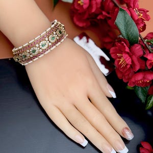 May include: An ornate gold-toned bracelet with red and green gemstones and small white pearls. The bracelet is on a mannequin hand with a French manicure. Red flowers are in the background.