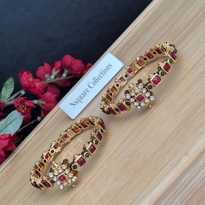 May include: Two gold-toned bangles with red and green gemstones. Each bangle features a central floral design with white gemstones. The bangles are displayed on a wooden tray with a white label that reads "Nsquare Collections".