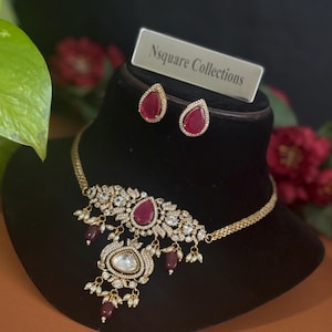 May include: A gold necklace with a teardrop-shaped pendant set with a large clear stone and smaller clear stones. The pendant is surrounded by smaller red stones. The necklace is accompanied by a pair of matching earrings with red stones. The card reads "Nsquare Collections".