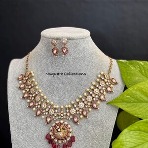 May include: A gold necklace with a peacock pendant and red stones. The necklace is adorned with white stones and pearls. The earrings are also gold with red and white stones.