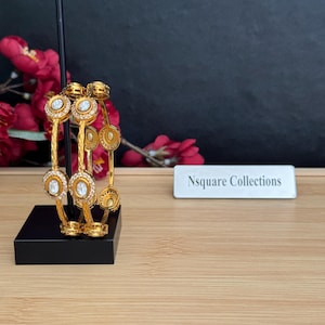 May include: Gold-toned bangles with clear, round, and oval-shaped embellishments. The bangles are displayed on a black stand. The text "Nsquare Collections" is on a white rectangular sign.