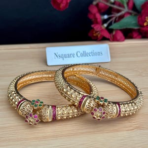 May include: Two gold-toned metal bangles featuring intricate textured patterns, a hinged opening, and decorative floral accents set with small red and green stones. A white rectangular sign with the text Nsquare Collections is visible.