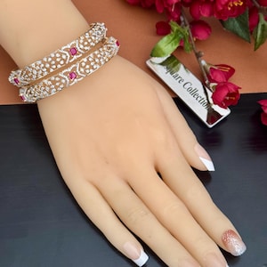 May include: Two ornate, silver-toned bangles with clear and red gemstones are displayed on a hand. The bangles feature a delicate, openwork design. The hand has a French manicure. A tag with the text "Square Collection" is visible.