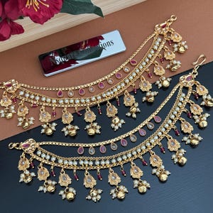 May include: A pair of gold-toned ear chains featuring multiple layered chains adorned with small white beads, teardrop-shaped red stones, and dangling bell-shaped charms. Each piece includes a hook for attachment and intricate metalwork details.