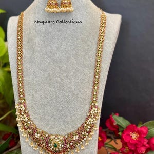May include: A gold-toned long necklace and matching jhumka earrings set featuring intricate filigree work, inlaid with red and green stones, white accents, and small dangling pearls. The text Nsquare Collections is visible.