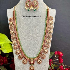 May include: A gold-toned necklace and matching earrings set. The necklace features a central pendant and circular accents with red and white details, complemented by green beads. The earrings have a similar design. The text "Nsquare Collections" is visible.