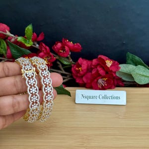 May include: Two gold-tone bangles featuring a repeating floral pattern encrusted with clear white stones. The bangles have a hinged clasp mechanism and are displayed alongside a white rectangular sign reading Nsquare Collections.