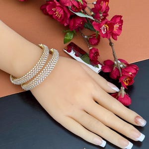 May include: Two gold-toned bangles encrusted with sparkling clear crystals. The bangles are on a hand with a French manicure and a glitter accent nail. Red flowers and a silver lipstick case are in the background.