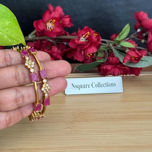 Könnte beinhalten: Goldfarbene Armreifen mit rosa rechteckigen und klaren runden Edelsteinen. Die Armreifen werden vor roten Blumen und einem Schild mit der Aufschrift "Nsquare Collections" präsentiert.