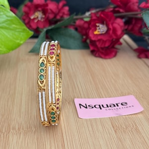 May include: Two gold bangles with white beads and green and red stones. The bangles are stacked on top of each other. The background is a wooden surface with a pink card that says "Nsquare Collections".