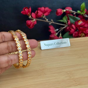 May include: Two gold-toned bangles featuring a repeating geometric pattern set with clear white stones and occasional small red and green accents. The bangles have a textured, matte finish and a structured, angular design along the outer edge.