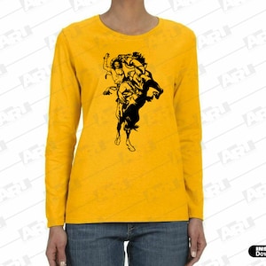 May include: A mustard yellow long-sleeve shirt featuring a black graphic print of a cowboy riding a bucking horse. The shirt has a crew neckline and is paired with blue denim jeans. The design depicts a rider in a hat on a rearing horse.