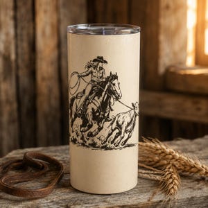 Op de afbeelding: Een cilindervormige beige beker met een zwarte lijntekening van een cowgirl op een paard die een kalf vangt met een lasso. Het ontwerp toont de ruiter met een lasso, waarbij het paard en het kalf in beweging zijn. De beker heeft een doorzichtig deksel.