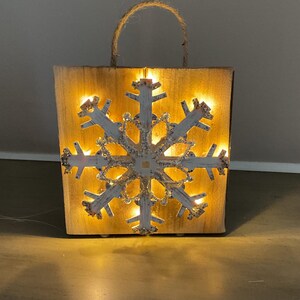 May include: A square wooden sign featuring a cutout snowflake design illuminated by warm yellow LED lights from behind. The natural wood grain is visible, and a jute rope handle is attached to the top edge for hanging or carrying purposes.