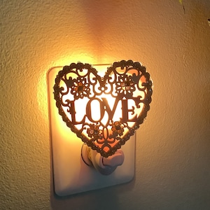 May include: A gold heart-shaped night light with the word "LOVE" cut out in the center. The light is plugged into an outlet and is glowing.