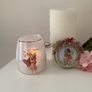 May include: A white candle, a glass votive candle holder with a fairy and the word "Merry" on it, and a round, gold-colored ornament with a fairy and the word "Merry" on it. The ornament is decorated with red berries and glitter.
