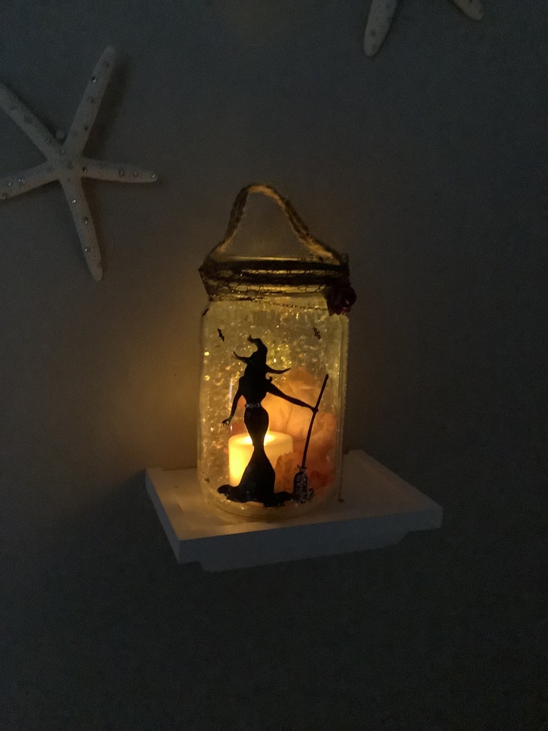 May include: A decorative glass jar with a witch silhouette, lit by a candle. The jar has a handle and is decorated with glitter and a rose. The witch is holding a broom. The jar is sitting on a white shelf.