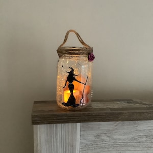 May include: A clear glass jar decorated with a witch silhouette, a broom, and glitter. The jar has a twine handle and a purple flower accent. Inside, a battery-operated candle glows, creating a warm, inviting Halloween decoration.