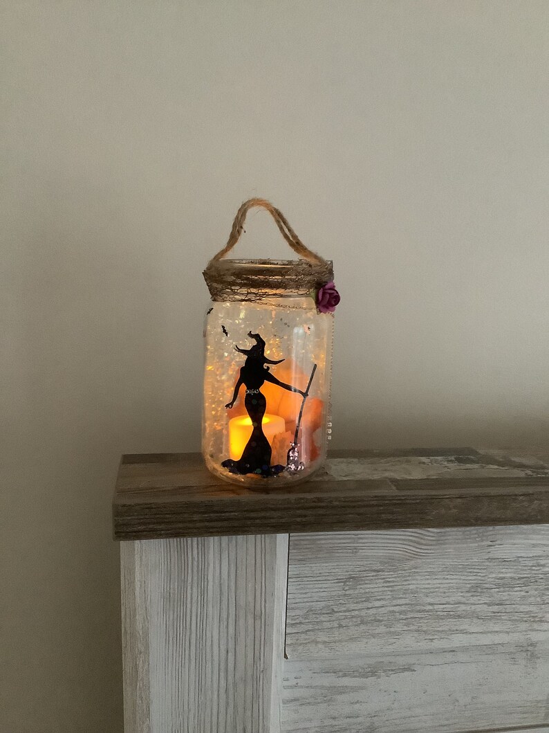May include: A clear glass jar decorated with a witch silhouette, a broom, and glitter. The jar has a twine handle and a purple flower accent. Inside, a battery-operated candle glows, creating a warm, inviting Halloween decoration.