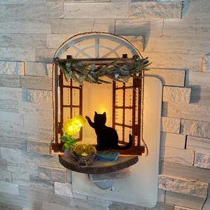 May include: A decorative night light shaped like a window with a black cat silhouette. The window features a small shelf with a bird's nest, a book, and a yellow flower. The light is plugged into a wall outlet.